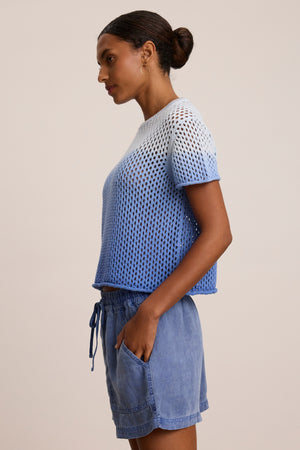 A person in profile wears the POPPY COTTON CASHMERE MESH SWEATER by Velvet by Graham & Spencer, featuring blue and white dip dye short sleeves, paired with light blue drawstring shorts, hands in pockets, against a plain background.