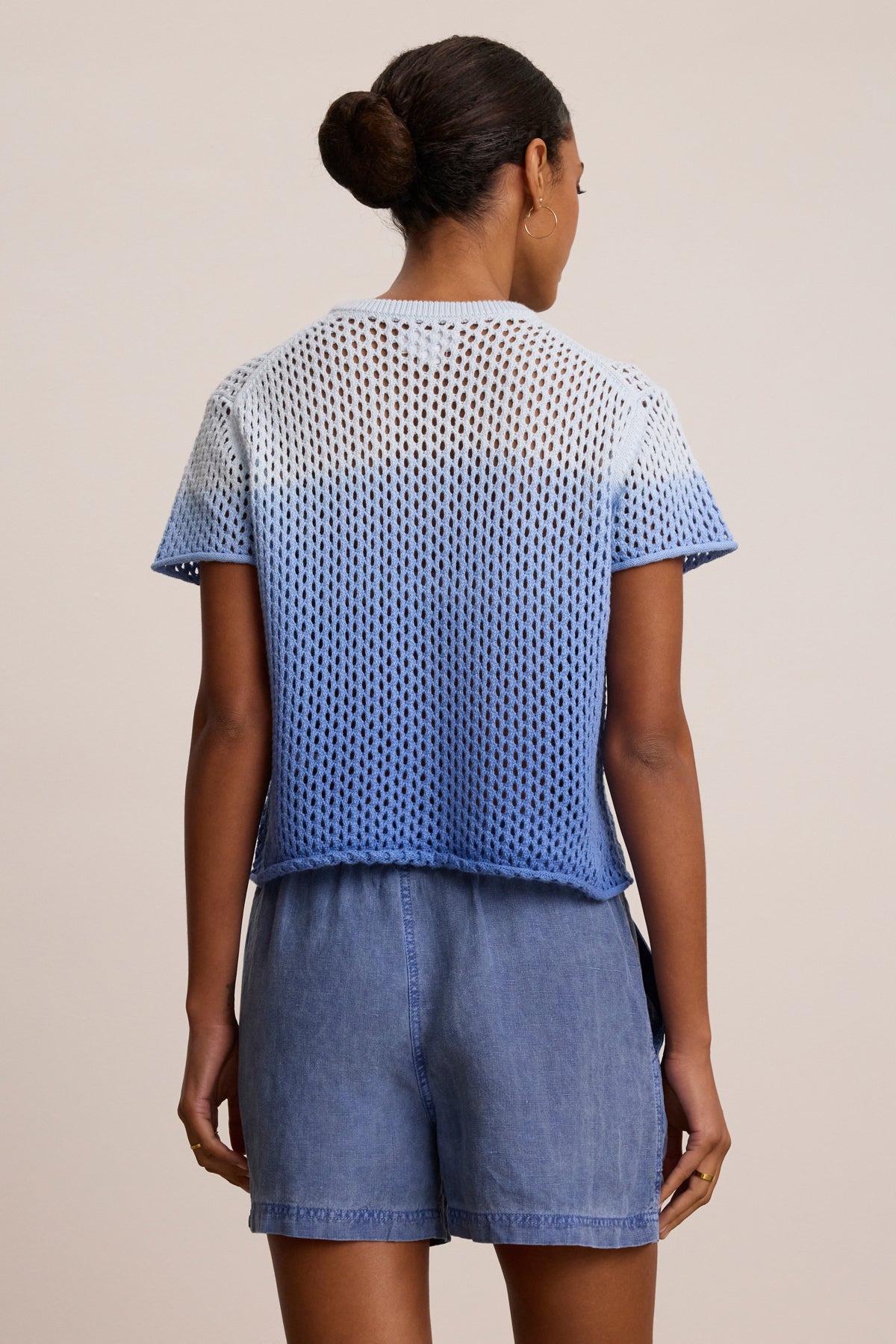 A woman faces away, wearing the POPPY Cotton Cashmere Mesh Sweater by Velvet by Graham & Spencer in blue and white ombre with light blue denim shorts, her hair styled in a neat bun.