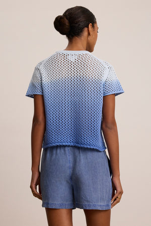 A woman faces away, wearing the POPPY Cotton Cashmere Mesh Sweater by Velvet by Graham & Spencer in blue and white ombre with light blue denim shorts, her hair styled in a neat bun.