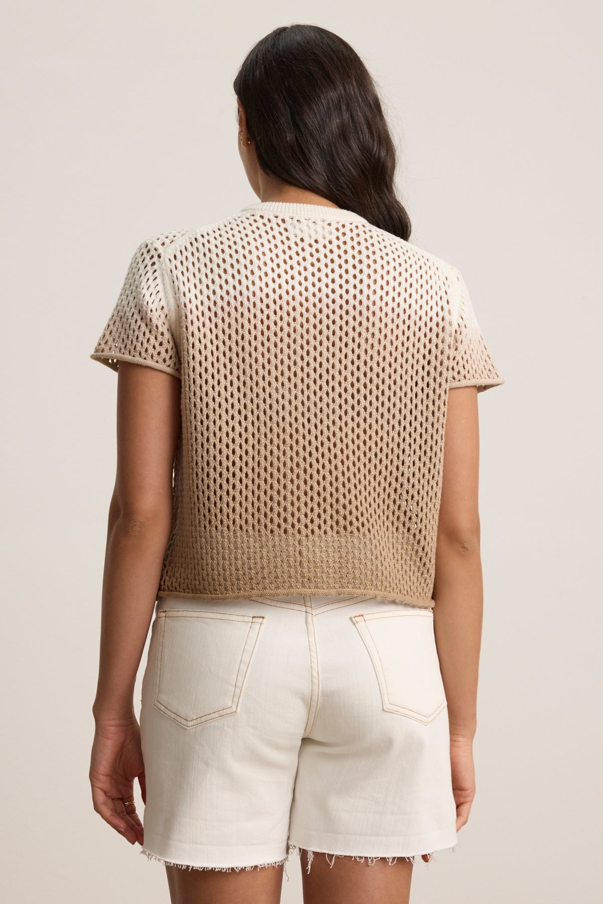 A woman stands with her back to the camera, wearing the Velvet by Graham & Spencer POPPY Cotton Cashmere Mesh Sweater in beige and white raw-hem denim shorts against a plain background.
