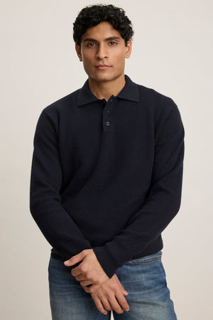 A man with short, curly dark hair wears the THALEN SWEATER POLO by Velvet by Graham & Spencer, a textured dark navy knit-collar sweater, paired with blue jeans, standing against a plain light background.