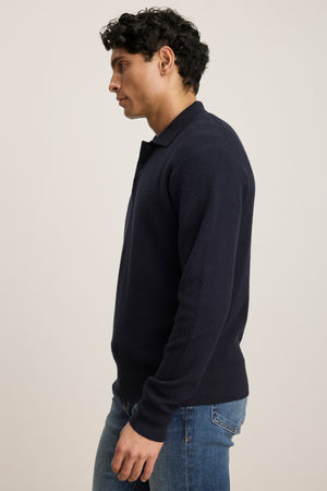 A man stands in profile wearing the Velvet by Graham & Spencer THALEN SWEATER POLO with a knit collar and blue jeans against a plain light background.