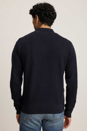 A man stands with his back to the camera against a plain light background, wearing the THALEN SWEATER POLO by Velvet by Graham & Spencer—a navy blue textured cotton cashmere blend sweater—paired with blue jeans.