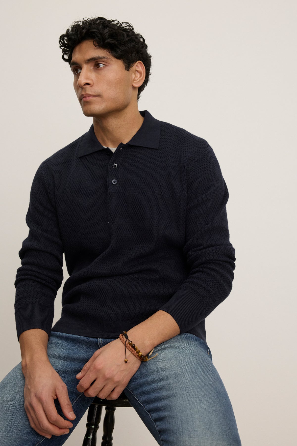 A man with short curly dark hair sits on a stool against a plain light background, wearing the THALEN SWEATER POLO by Velvet by Graham & Spencer and blue jeans.