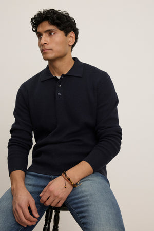 A man with short curly dark hair sits on a stool against a plain light background, wearing the THALEN SWEATER POLO by Velvet by Graham & Spencer and blue jeans.