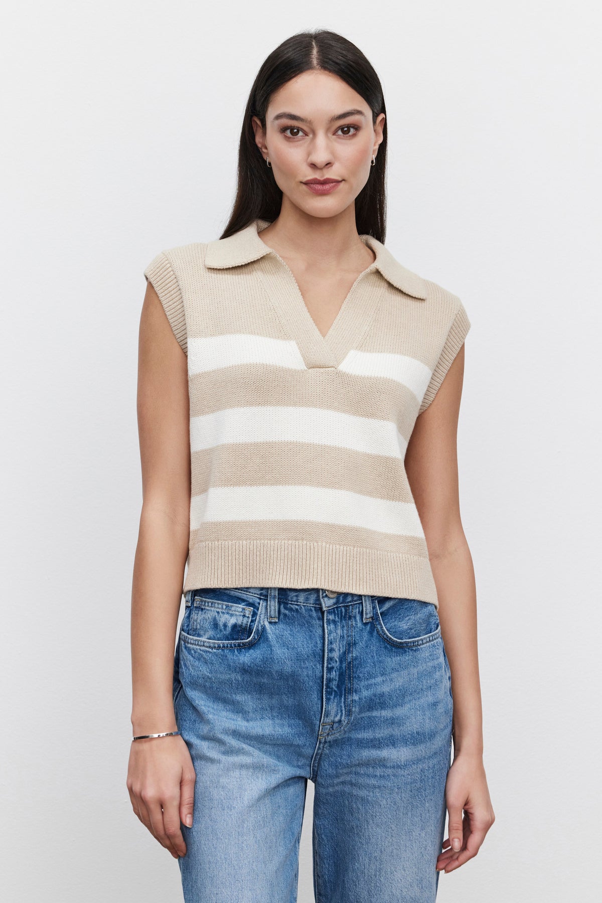 A woman with long hair wears the MARIETTE SWEATER VEST, a beige and white striped sleeveless knit top from Velvet by Graham & Spencer, made of soft cotton. She pairs it with blue jeans as she stands against a plain white background, looking directly at the camera.-37629552394433