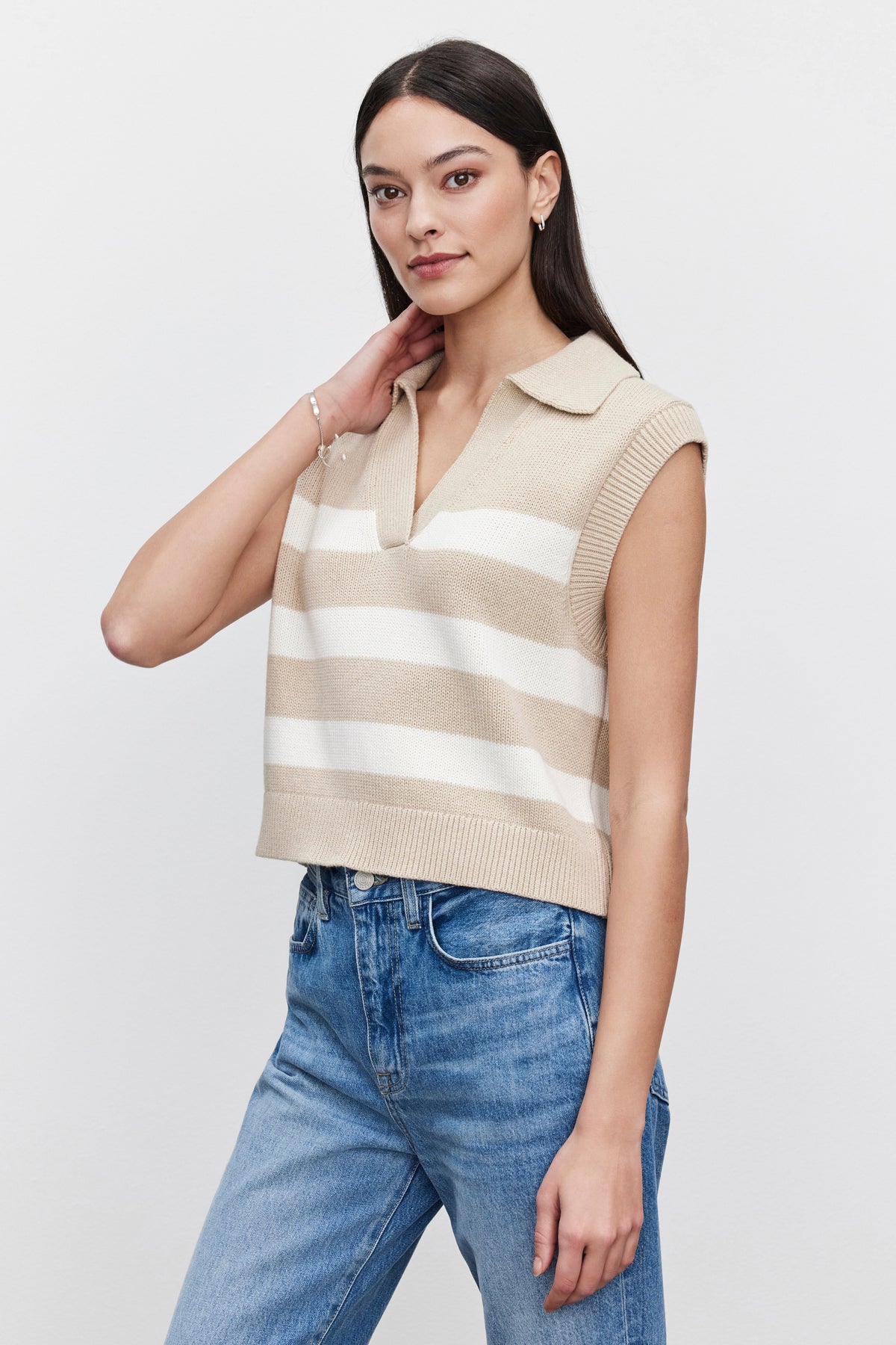 A woman with long dark hair wears the MARIETTE SWEATER VEST by Velvet by Graham & Spencer paired with jeans. She poses against a plain white background, one hand touching her hair.