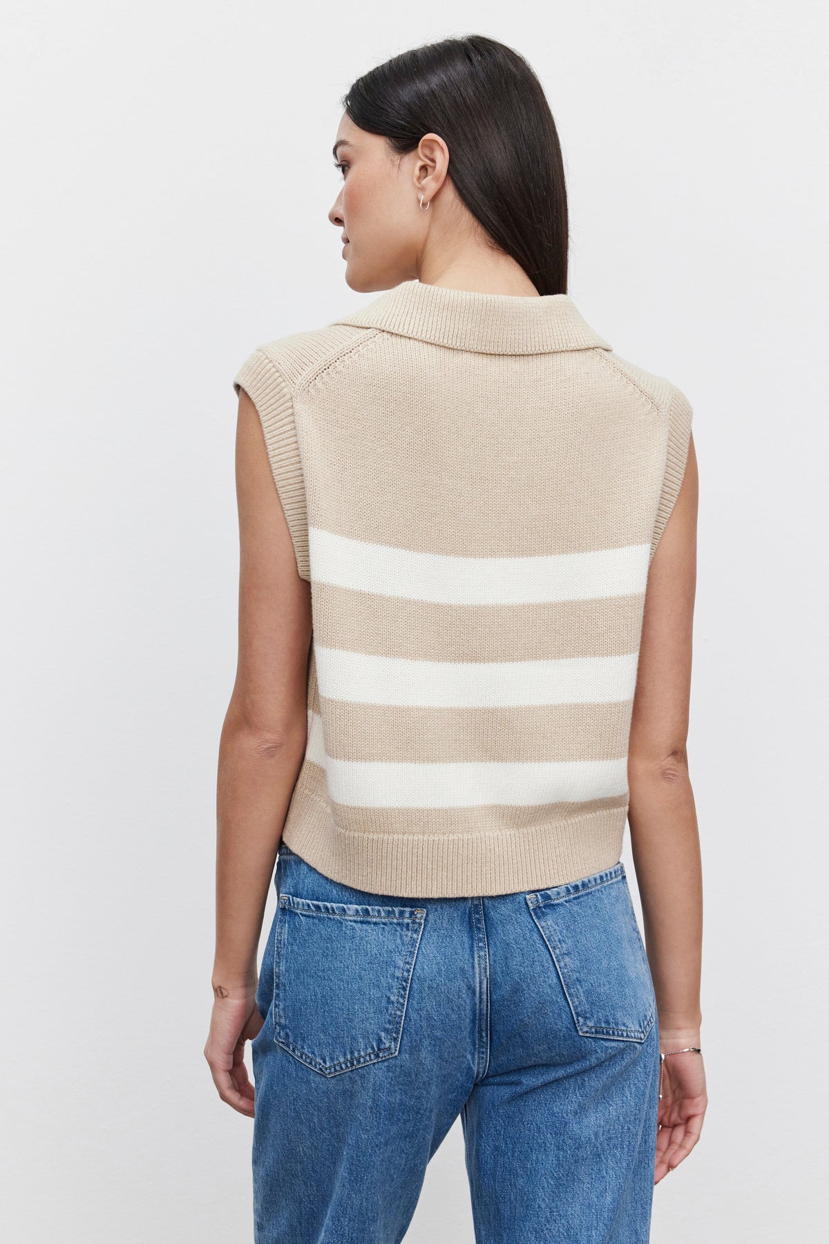 A woman with long dark hair, seen from behind and standing against a plain white background, is wearing the MARIETTE SWEATER VEST by Velvet by Graham & Spencer, which is a sleeveless beige cashmere sweater with white stripes on the back, paired with blue jeans.