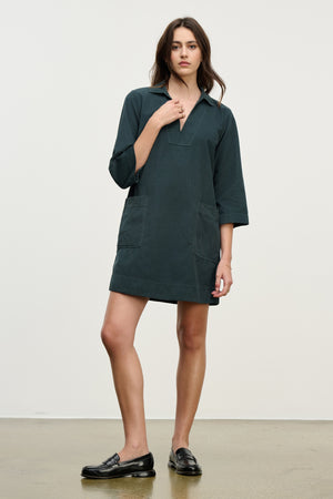 A woman stands indoors wearing the ELKE CORDUROY DRESS by Velvet by Graham & Spencer—a dark green, long-sleeve, collared cotton dress with front pockets—paired with black loafers for effortlessly stylish everyday wear.