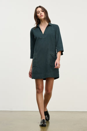 A woman stands against a plain background, wearing the ELKE CORDUROY DRESS by Velvet by Graham & Spencer—a dark green, long-sleeve cotton shift dress with front pockets, ideal for everyday wear.