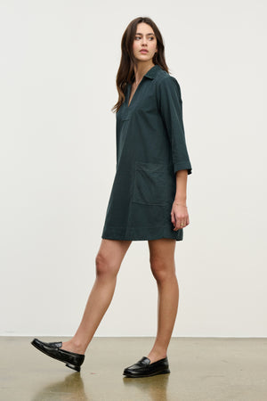 A woman in the ELKE CORDUROY DRESS by Velvet by Graham & Spencer—dark green, long-sleeve cotton with front pockets—pairs it with black loafers on a white backdrop, making it perfect for everyday wear.