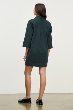 A woman faces away in the ELKE CORDUROY DRESS by Velvet by Graham & Spencer, a dark green cotton mini dress with three-quarter sleeves, paired with black loafers against a plain background.