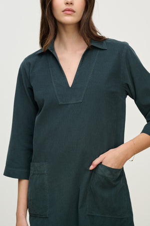 A woman wears the ELKE CORDUROY DRESS by Velvet by Graham & Spencer—a dark green cotton corduroy V-neck mini dress with three-quarter sleeves and large front pockets, perfect for everyday wear.