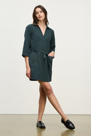 A woman stands against a plain background wearing the ELKE CORDUROY DRESS by Velvet by Graham & Spencer—A dark green, belted cotton mini dress with three-quarter sleeves, styled with black loafers for effortless everyday wear.