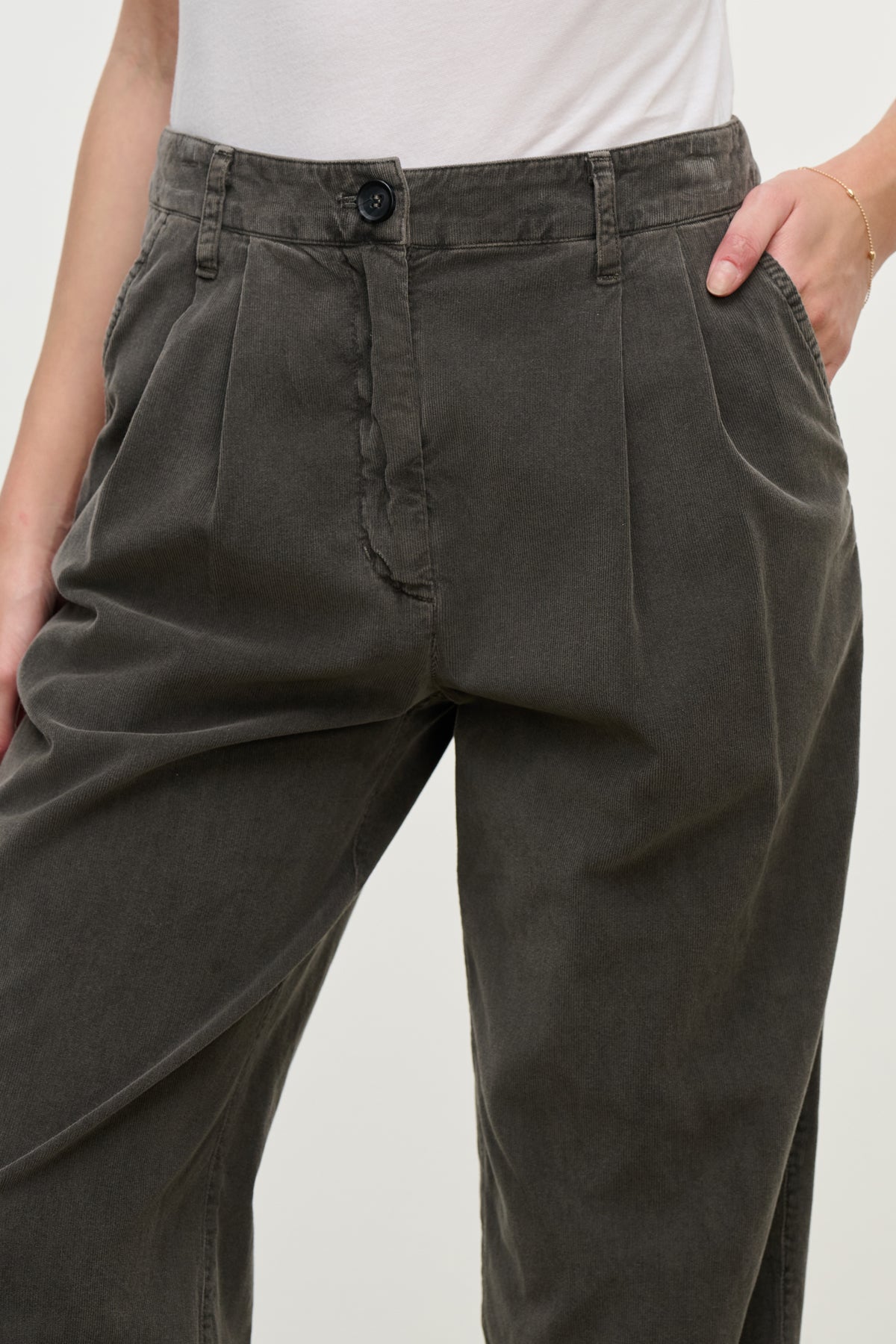 Model wearing Velvet by Graham & Spencer's GAIL CORDUROY PANT, a high-waisted, loose-fitting dark gray pleated straight-leg style, paired with a white shirt and hands in pockets. Shown from waist to mid-thigh.
