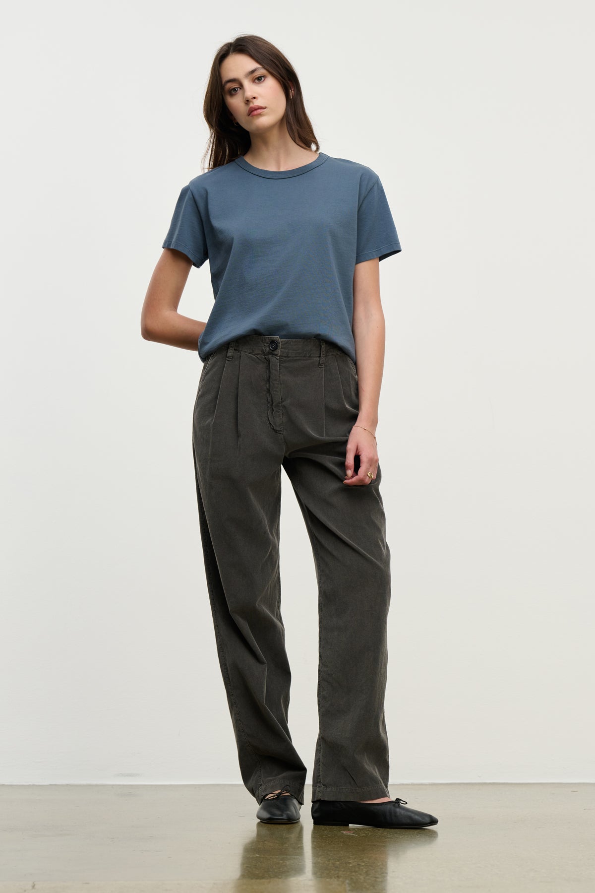 A person stands against a plain background wearing the GAIL CORDUROY PANT by Velvet by Graham & Spencer in dark cotton, paired with a blue t-shirt and black shoes, with one hand behind their back.