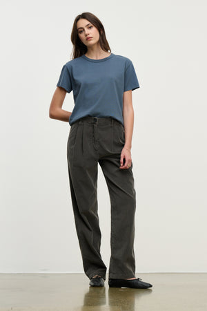 A person stands against a plain background wearing the GAIL CORDUROY PANT by Velvet by Graham & Spencer in dark cotton, paired with a blue t-shirt and black shoes, with one hand behind their back.