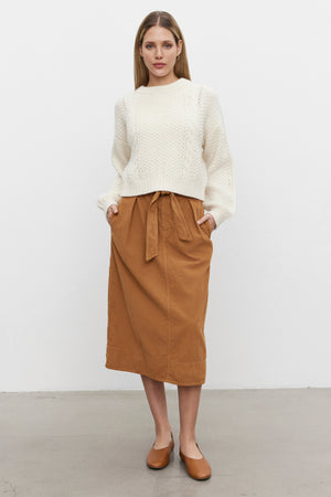 A person stands indoors wearing a white knit sweater, the Velvet by Graham & Spencer KENNEDY CORDUROY SKIRT with a detachable belt, and brown shoes. They have their hands in their skirt pockets and are looking towards the camera.