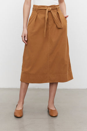 A woman stands against a plain background, wearing a KENNEDY CORDUROY SKIRT by Velvet by Graham & Spencer, a tan cotton corduroy midi skirt with a waist tie, paired with matching tan shoes.