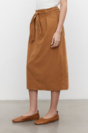 A person wearing a white top and the KENNEDY CORDUROY SKIRT by Velvet by Graham & Spencer, paired with brown flats. Only the lower part of the body is visible.