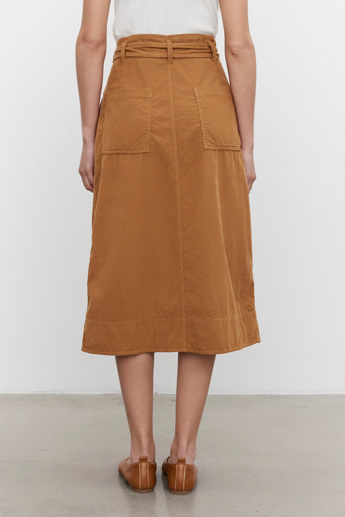 A person wearing a white top and the KENNEDY CORDUROY SKIRT by Velvet by Graham & Spencer, a brown cotton corduroy midi skirt with front pockets, photographed from the back, standing on a gray floor against a plain white background.-37856324747457