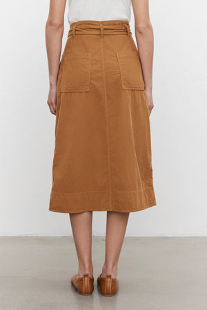 A person wearing a white top and the KENNEDY CORDUROY SKIRT by Velvet by Graham & Spencer, a brown cotton corduroy midi skirt with front pockets, photographed from the back, standing on a gray floor against a plain white background.