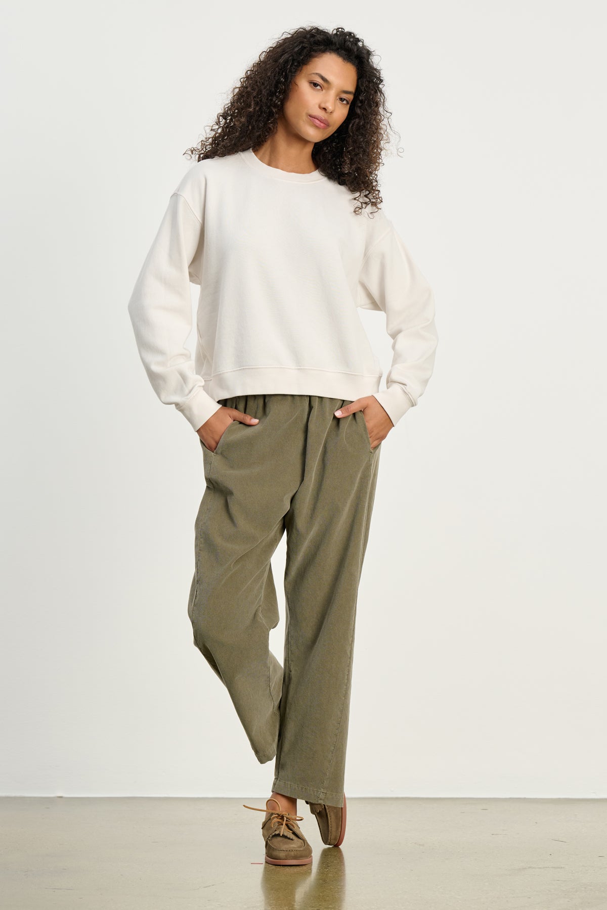 A person stands with hands in pockets, wearing a white sweatshirt and Velvet by Graham & Spencer's LANEY CORDUROY PANT in olive green with an elastic pull-on waist, plus brown shoes, against a plain white background.-39539207930049