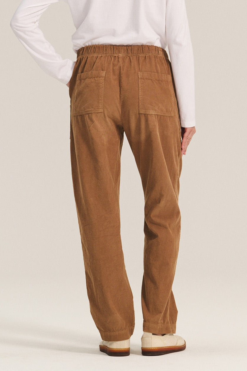 A person faces away, wearing Velvet by Graham & Spencer's ASPEN CORDUROY PANT in brown with an elastic waist and rear pockets, plus a white long-sleeve shirt and slip-on shoes, against a plain light background.-40248297029825