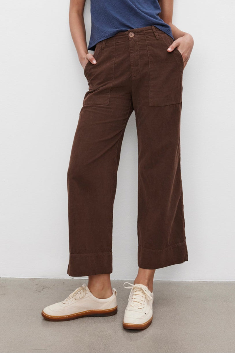 A person stands against a plain background wearing the MARIE TEE by Velvet by Graham & Spencer, a blue cotton slub t-shirt with a relaxed boxy fit, paired with brown wide-leg pants and hands in pockets, and white sneakers—perfect for a casual occasion.