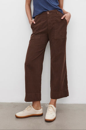 A person stands against a plain background wearing the MARIE TEE by Velvet by Graham & Spencer, a blue cotton slub t-shirt with a relaxed boxy fit, paired with brown wide-leg pants and hands in pockets, and white sneakers—perfect for a casual occasion.