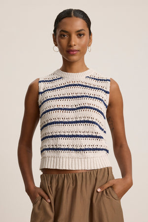 A person wears the CALISTA SWEATER VEST by Velvet by Graham & Spencer—a sleeveless, knit cream cropped sweater vest with navy and black stripes—paired with brown high-waisted pants, standing hands in pockets against a plain background.