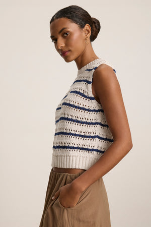 A woman in profile wears the CALISTA SWEATER VEST by Velvet by Graham & Spencer, featuring blue and white stripes, paired with light brown pants; her hair is pulled back in a low bun.