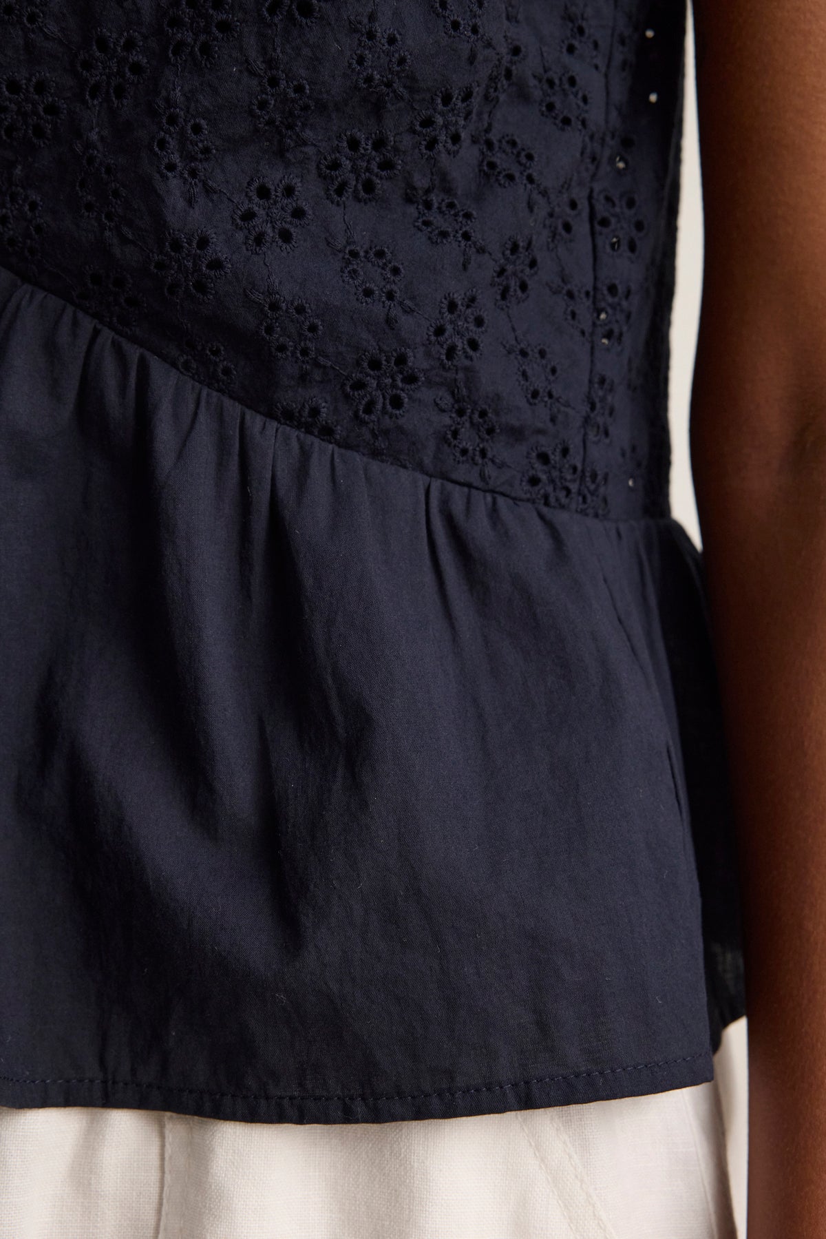 Close-up of a person wearing the ALIAH EYELET TANK TOP by Velvet by Graham & Spencer, featuring navy blue eyelet detailing, a tiered silhouette, and ruffled hem, paired with white bottoms.