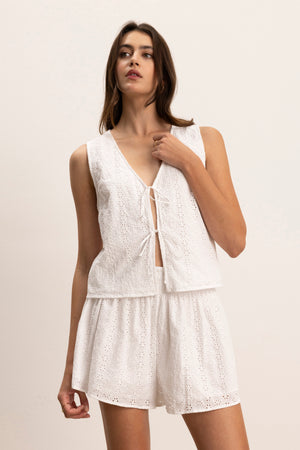 A woman wears the AVA EYELET TOP by Velvet by Graham & Spencer—a sleeveless, white cotton V-neck with front tie—paired with matching eyelet shorts, standing against a plain light background.