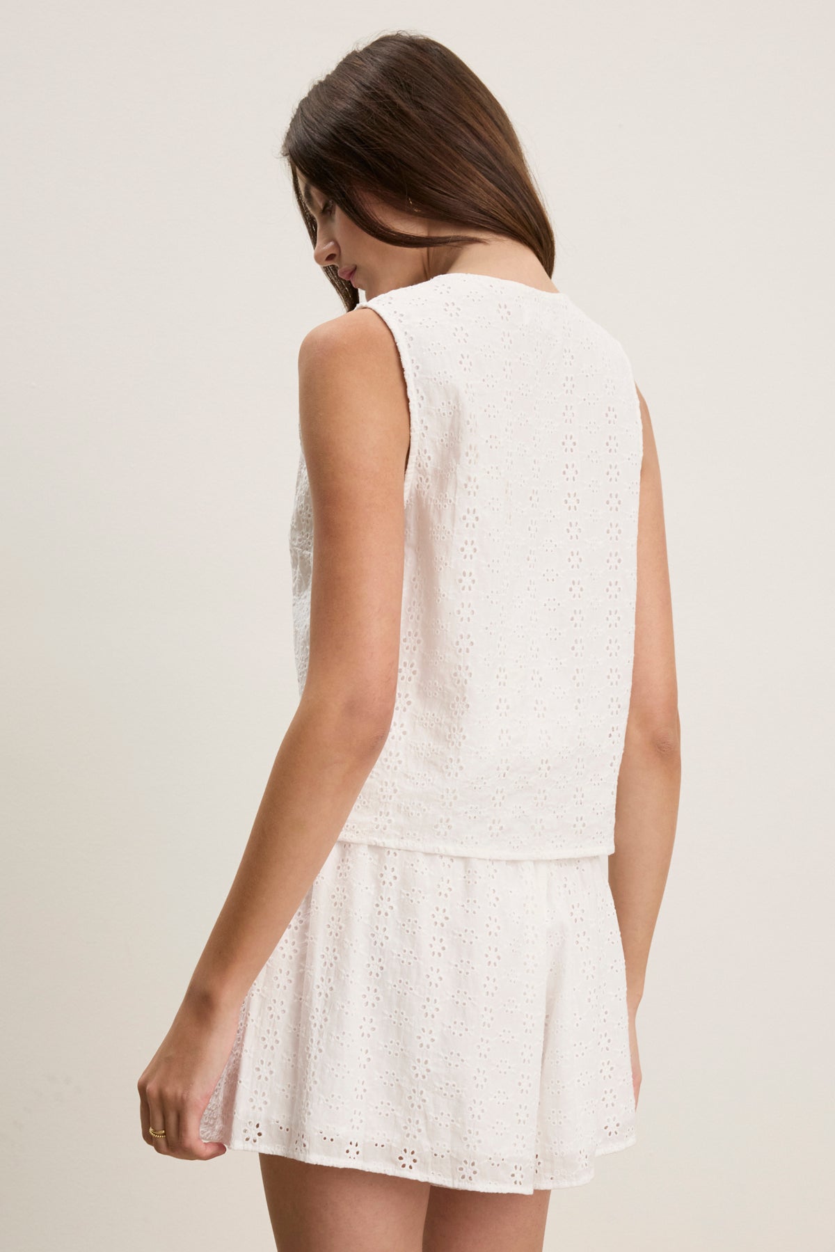 A woman faces away, wearing the AVA EYELET TOP by Velvet by Graham & Spencer, paired with a matching skirt. The sleeveless cotton design adds a feminine touch and versatility to her wardrobe against a plain backdrop.-39123271057601