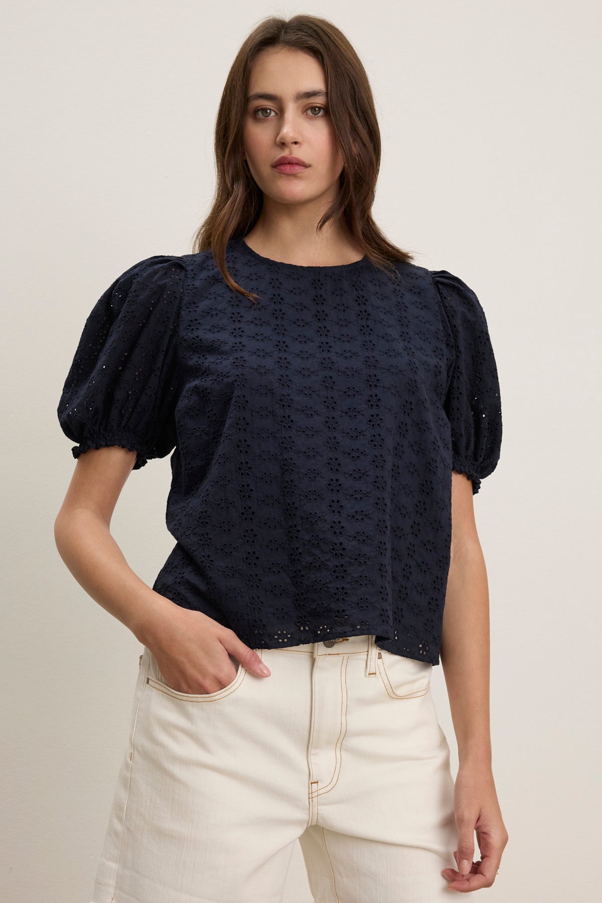 A woman stands against a plain background wearing the DALYN EYELET TOP by Velvet by Graham & Spencer—a navy cotton blouse with puff sleeves—and off-white jeans, with one hand in her pocket.-39123283869889