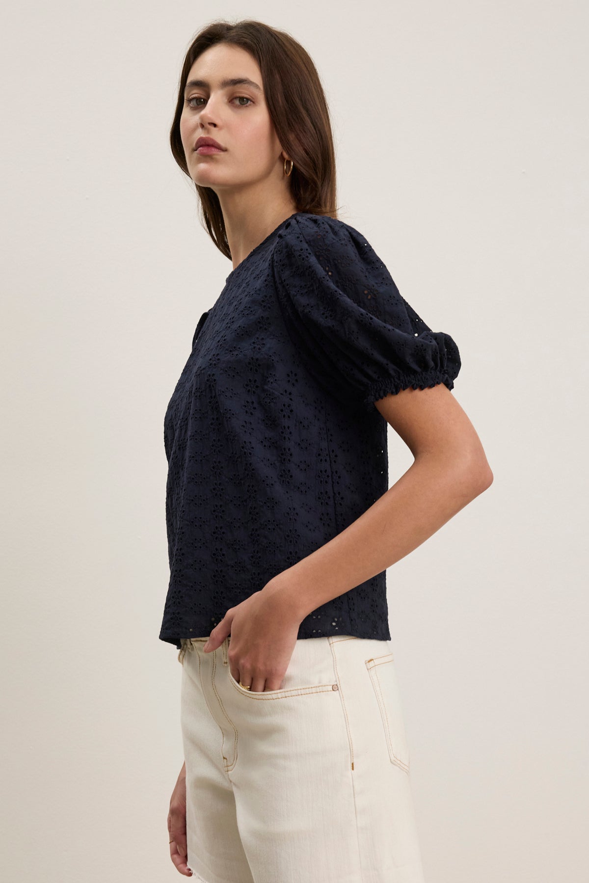 A woman stands against a plain background wearing the DALYN EYELET TOP by Velvet by Graham & Spencer, a navy cotton blouse with short puff sleeves, paired with high-waisted off-white jeans.-39123283837121