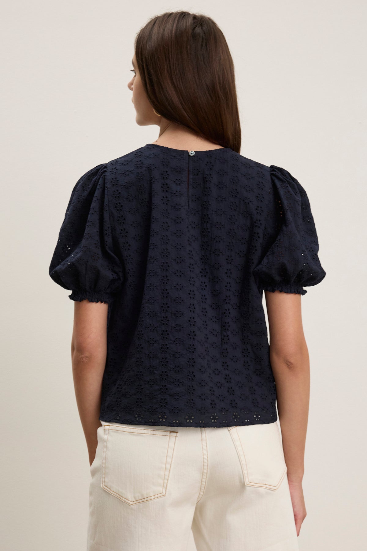 A woman faces away, wearing the DALYN EYELET TOP by Velvet by Graham & Spencer—a navy blue cotton blouse with short sleeves—paired with white pants against a plain light background.-39123283804353
