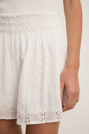 Someone is wearing the IRIS EYELET SHORT skirt by Velvet by Graham & Spencer in white, featuring a comfortable fit and elastic waistband, paired with a white top and showing their right hand with a ring.