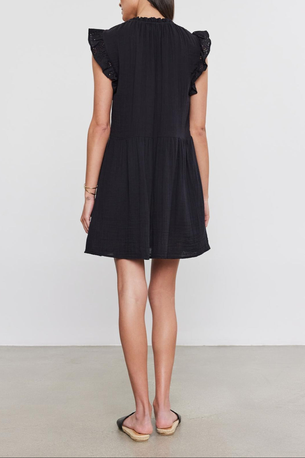 A woman in a black Velvet by Graham & Spencer dress with cotton eyelet flutter sleeves stands facing away, on a plain light background.-36910266253505