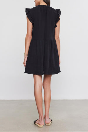 A woman in a black Velvet by Graham & Spencer dress with cotton eyelet flutter sleeves stands facing away, on a plain light background.