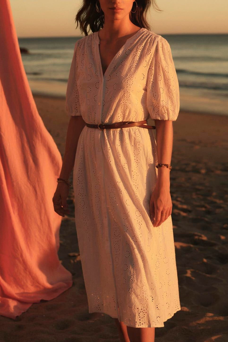 A woman in a white cotton eyelet RORI DRESS from Velvet by Graham & Spencer stands on a beach at sunset, with a pink fabric billowing in the background.-36910268055745