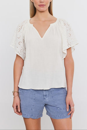 Woman in a white embroidered eyelet TISH TOP by Velvet by Graham & Spencer and blue denim shorts standing against a plain background.