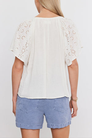 Rear view of a woman wearing a white TISH TOP by Velvet by Graham & Spencer with embroidered eyelet sleeves and blue denim shorts, standing against a white background.