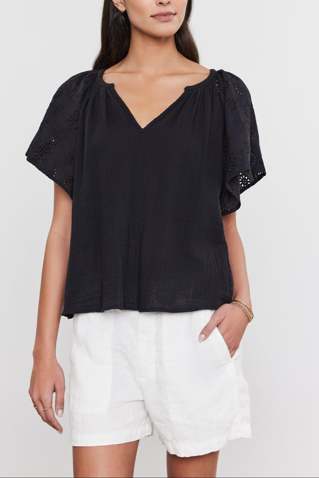 A woman wearing a black v-neck TISH TOP with embroidered eyelet sleeves and white shorts, standing against a white background, by Velvet by Graham & Spencer.-36918792945857