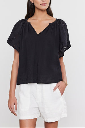 A woman wearing a black v-neck TISH TOP with embroidered eyelet sleeves and white shorts, standing against a white background, by Velvet by Graham & Spencer.