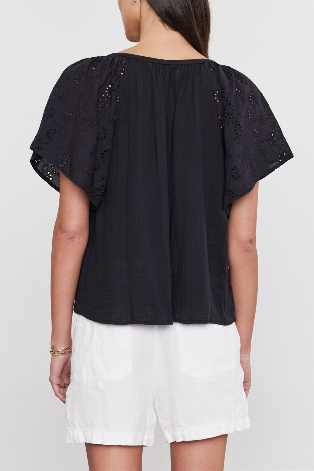 Woman standing with her back to the camera, wearing a black TISH TOP by Velvet by Graham & Spencer with embroidered eyelet details on the sleeves and white shorts.-36918793011393