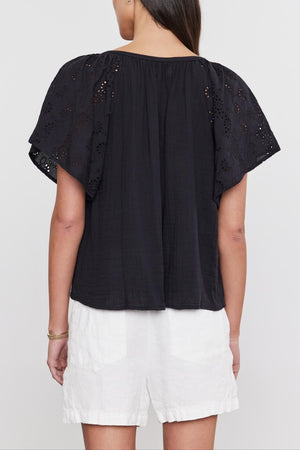 Woman standing with her back to the camera, wearing a black TISH TOP by Velvet by Graham & Spencer with embroidered eyelet details on the sleeves and white shorts.