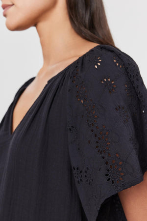 Close-up of a woman's shoulder and upper arm wearing a black TISH TOP blouse by Velvet by Graham & Spencer with intricate embroidered eyelet patterns.