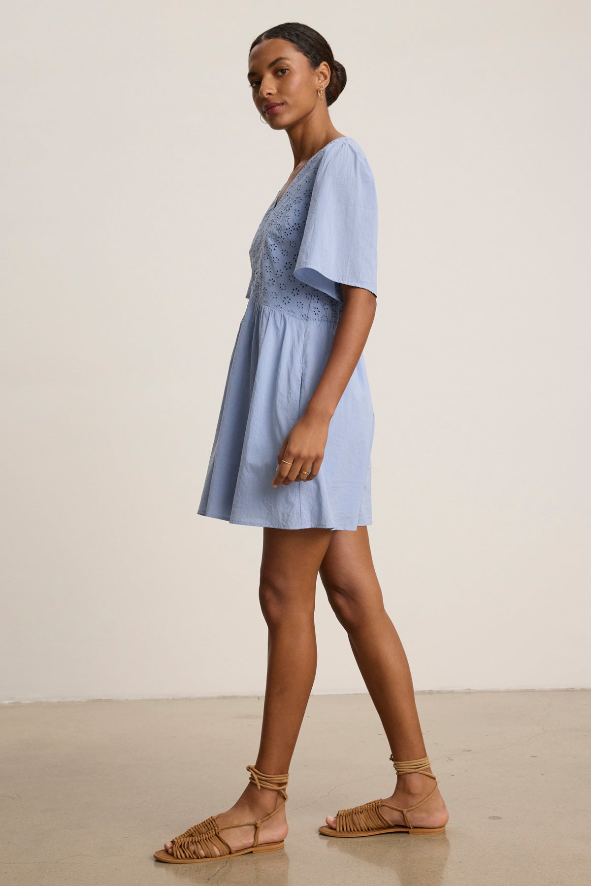 A woman stands sideways wearing the NELLI EYELET DRESS by Velvet by Graham & Spencer—a light blue cotton dress with short sleeves, V neckline, and fitted waist—paired with tan lace-up sandals against a plain light background.-39123389087937